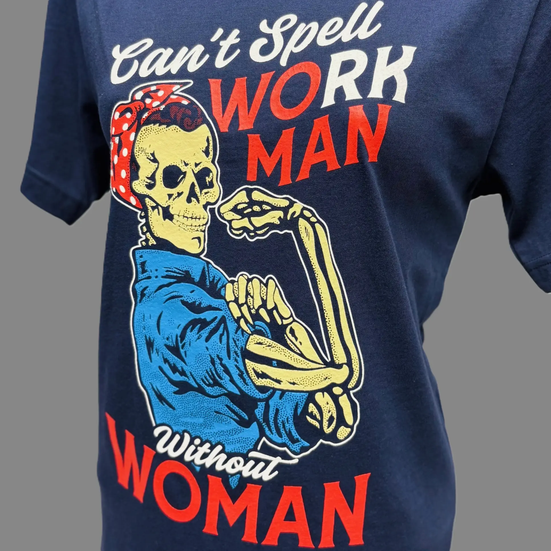 Navy blue shirt with text "Can't Spell workman without woman" "Can't Spell and Without are in white script. "Work Man" is in all caps with the WO and MAN in red text and the RK in white text. Woman is in all caps red text at the bottom. There is a a skeleton torso up (no legs) with a blue jumpsuit on and a red headband with white polka dots posing like Rosie the riveter.