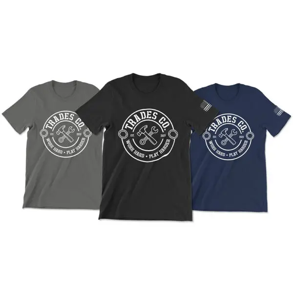 3 t-shirts, one black, one navy blue, one grey, with large Trades Co. Logo on the front center in white. Logo is a circular design with "TRADES CO" at the top and "WORK HARD ● PLAY HARDER" at the bottom with bolts centered on the outer center circle between the "trades co" and "work hard play harder". The center of the circle has a hammer and wrench crossed with est. on one side and 2023 on the other. White worn flag featured on left sleeve.