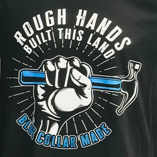Black t-shirt with white text in circular shape "Rough hands" with "Built this land" directly below it at the top of the circle with "Blue collar made" in blue text with black and white outline at the bottom of the circle. A white and black fist gripping a hammer with a blue handle emerges from the bottom text into the center of the circle shape. White lines come from all around the fist moving toward the outside of the circle.